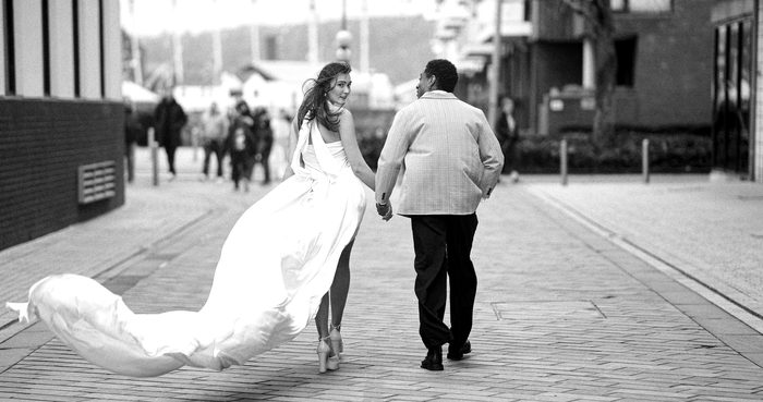 Black and white photo of a bride and groom walking hand in hand down a city street, her long dress trailing in the wind