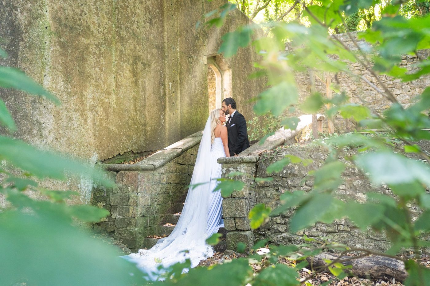 Bride and groom share a forehead kiss on moss-covered stone steps, framed by woodland leaves, photographed by Pedro de Sousa
