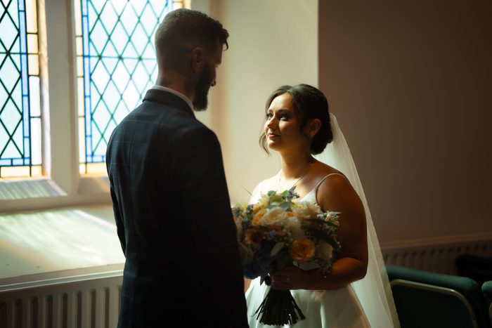 Bride holding her bouquet smiles up at the groom beside a leaded church window