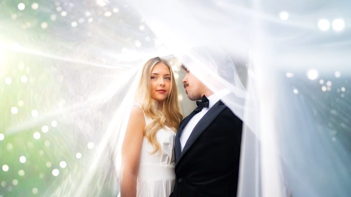 Bride and groom stand close beneath her veil, sunlight and sparkles of light flaring around them
