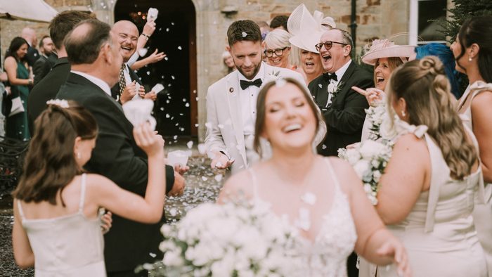 Bride laughing as guests throw confetti around her, the groom in a white tuxedo just behind