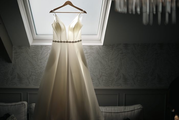 Satin wedding dress with a beaded belt hanging from a wooden hanger in front of a bright skylight