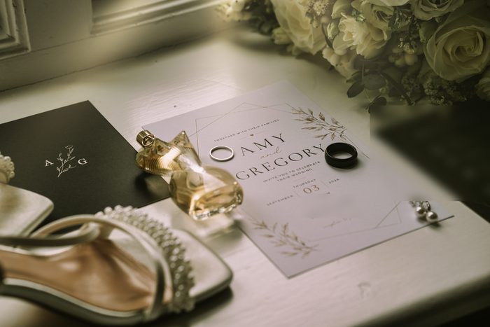 Wedding invitation with both rings, perfume, pearl earrings and pearl-strap bridal sandals laid out on a window sill beside the bouquet