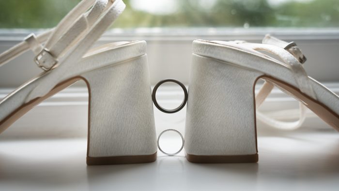 Two wedding rings balanced between the heels of a pair of ivory bridal shoes on a window sill